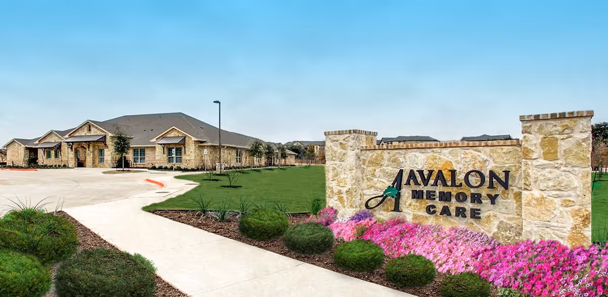 Exterior view of Avalon Memory Care facility showing a stone sign with the facility name surrounded by pink flowers and greenery, with a large building and clear blue sky in the background.