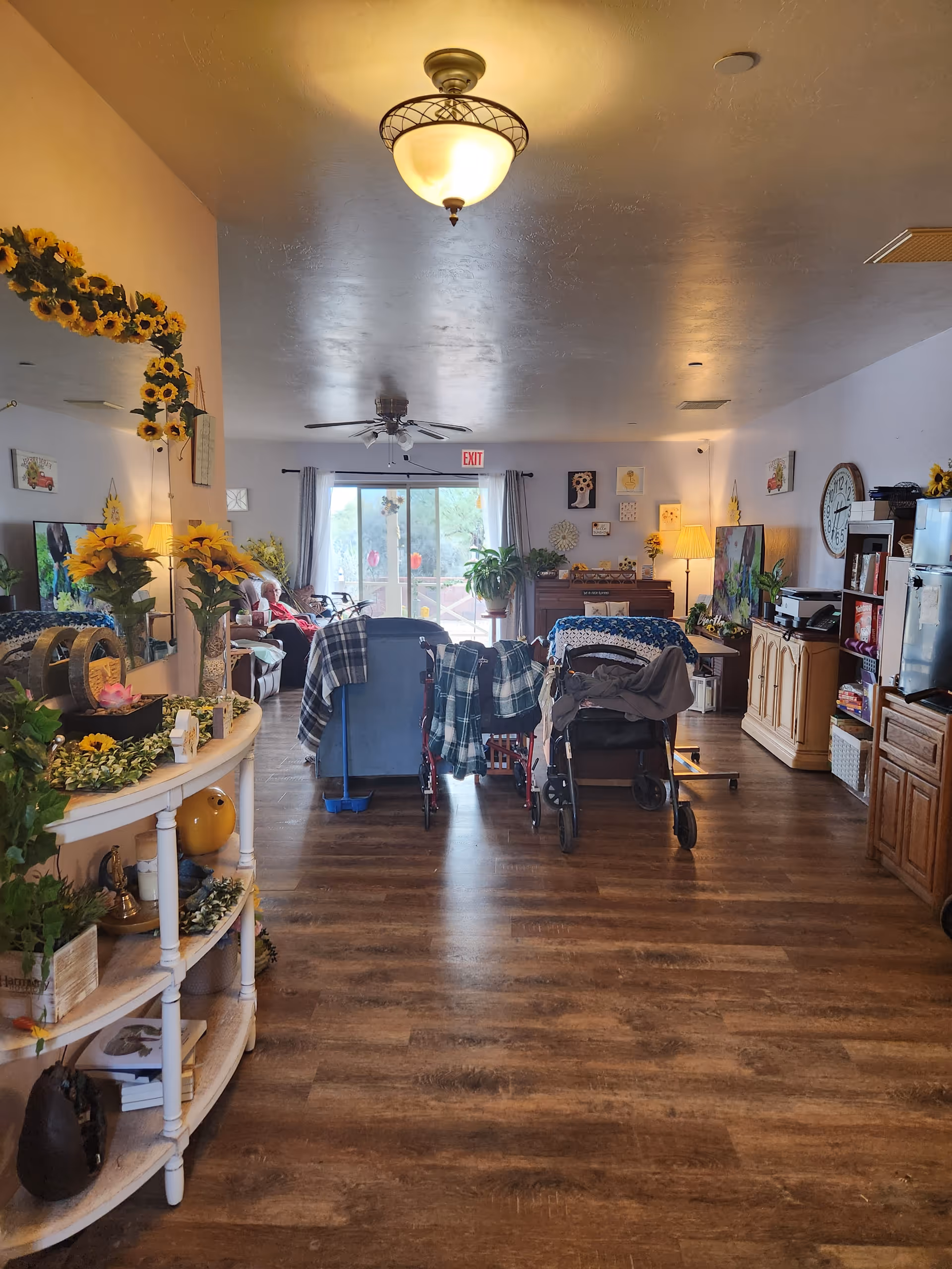 Interior view of a cozy assisted living common area with wooden flooring, decorated with sunflowers and various plants. There are chairs and wheelchairs arranged facing a large window with curtains, allowing natural light to enter. The room has a ceiling fan, a lamp, a clock on the wall, and shelves with books and decorative items.