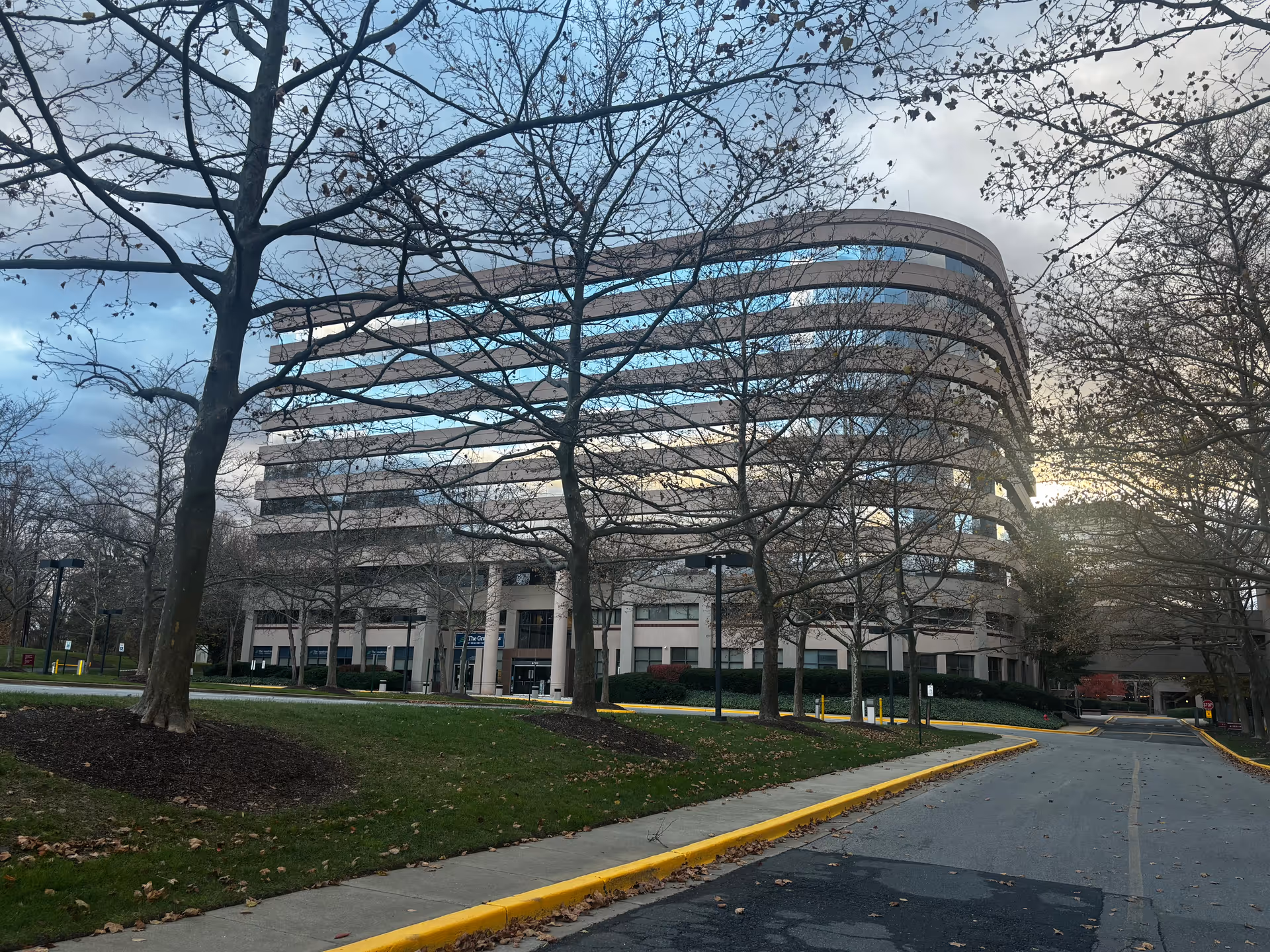 Curved multi-story office-style building behind leafless trees with a driveway and lawn in the foreground.