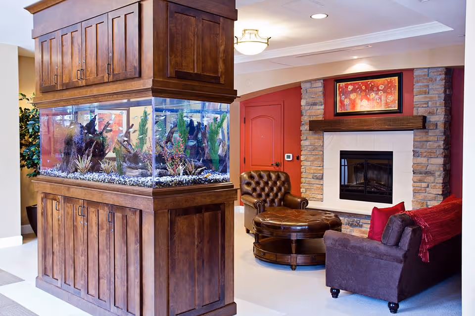 A cozy living room area featuring a large wooden aquarium filled with fish and aquatic plants. The room includes a brown leather armchair, a matching round ottoman, a dark brown sofa with red pillows and a red throw, and a stone fireplace with a wooden mantel. Above the fireplace hangs a colorful framed painting. The walls are painted in warm tones, including a red accent wall behind the fireplace.