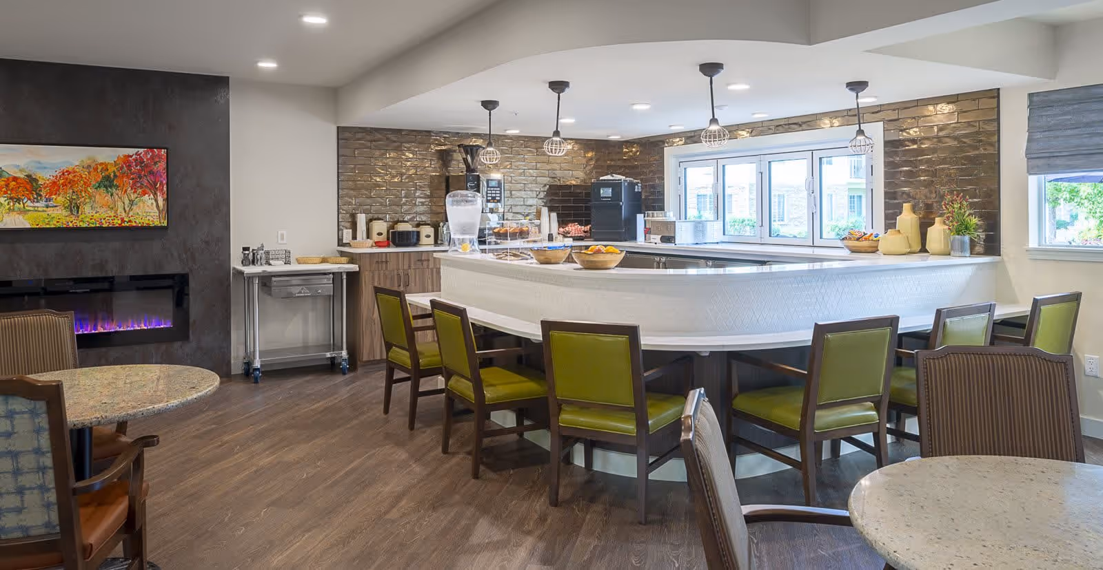 A modern senior living common area featuring a large U-shaped counter with green cushioned chairs around it. The counter has bowls of fruit and a water dispenser. Behind the counter is a kitchen area with appliances and a window. The room has wood flooring, a wall-mounted electric fireplace with a colorful painting above it, and additional round tables with chairs.