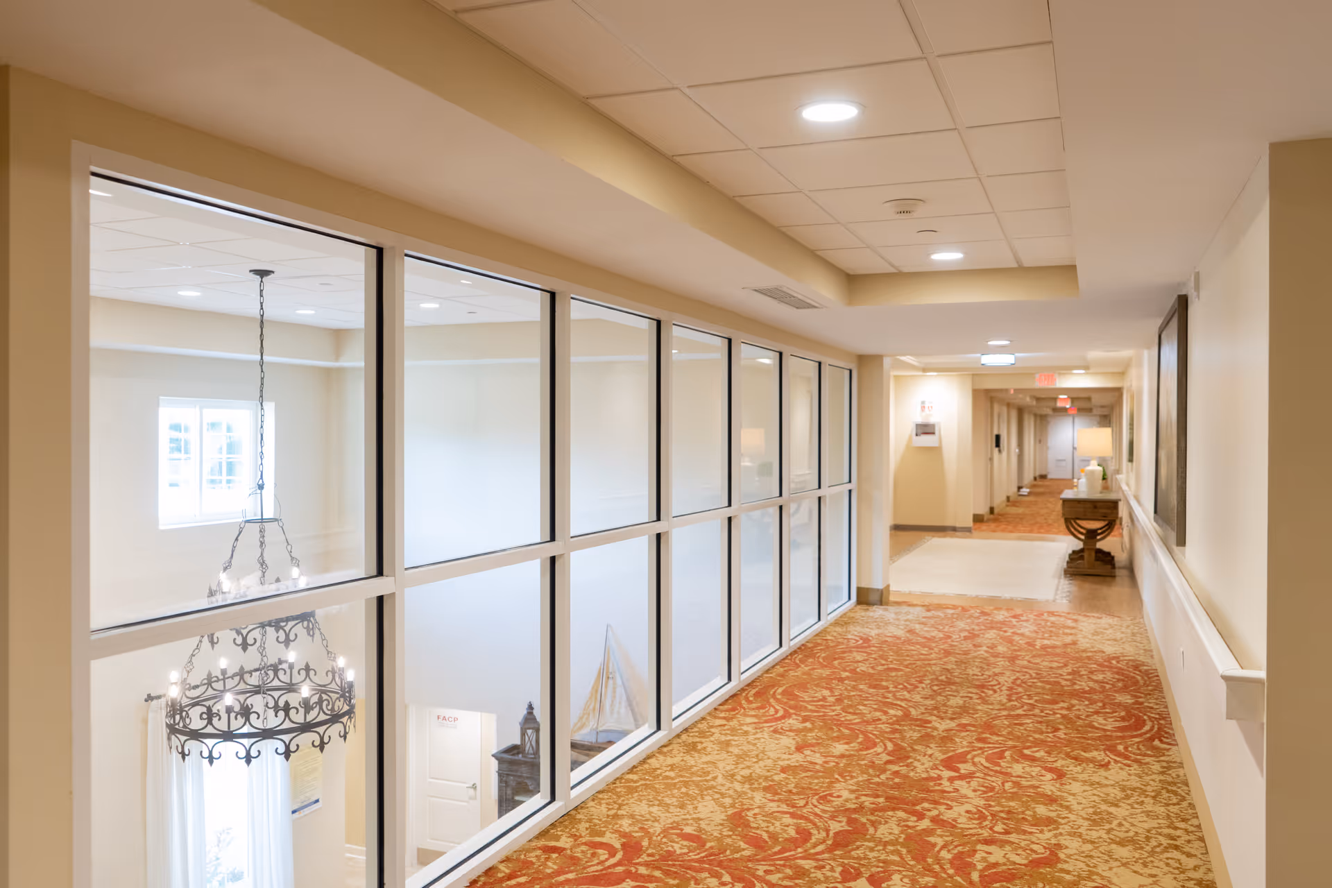 A well-lit interior hallway with a patterned red and beige carpet. On the left side, there is a large glass window wall overlooking a lower level with a decorative chandelier and a model sailboat. The hallway has beige walls, ceiling lights, and a small table with a lamp and decorative items on the right side.