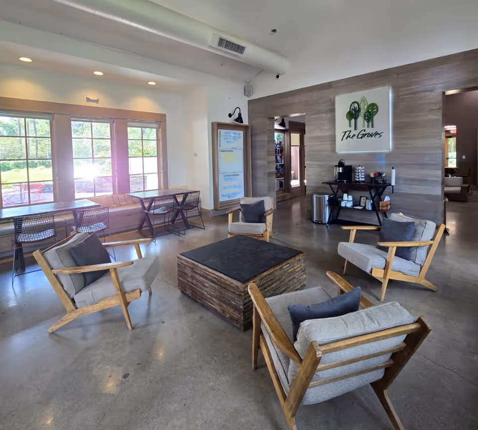 Bright communal lounge with wooden armchairs around a low square coffee table, large windows, and a coffee station beneath a 'The Groves' sign.