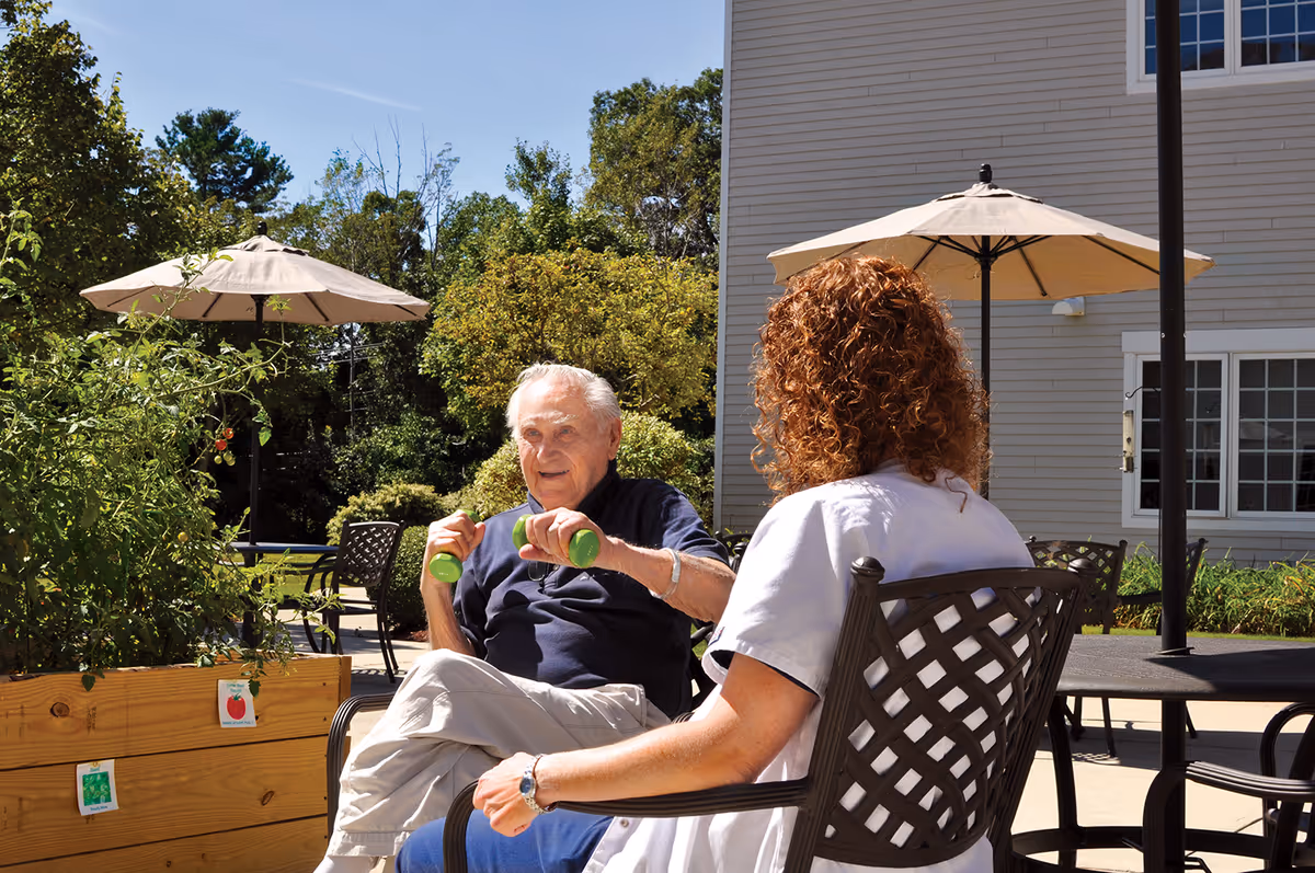An elderly man exercising with small green dumbbells while sitting outdoors at a table with a woman in a white uniform. They are in a garden area with plants, patio umbrellas, and outdoor furniture, next to a building under a clear blue sky.