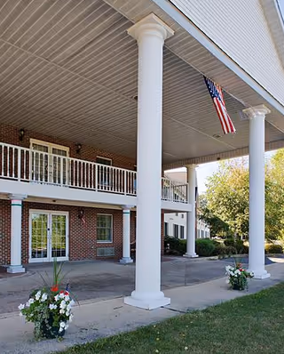 Exterior view of a brick senior living facility with a covered entrance, columns, and a flag.