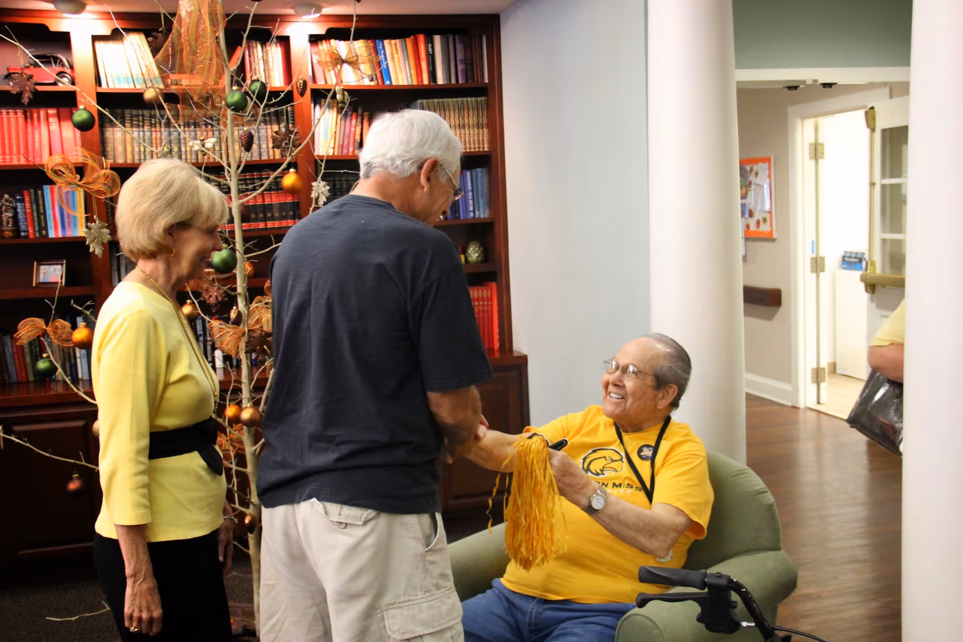 Three seniors greeting each other in a decorated common room with bookshelves; a seated man in a yellow shirt smiles and holds a yellow pom-pom.