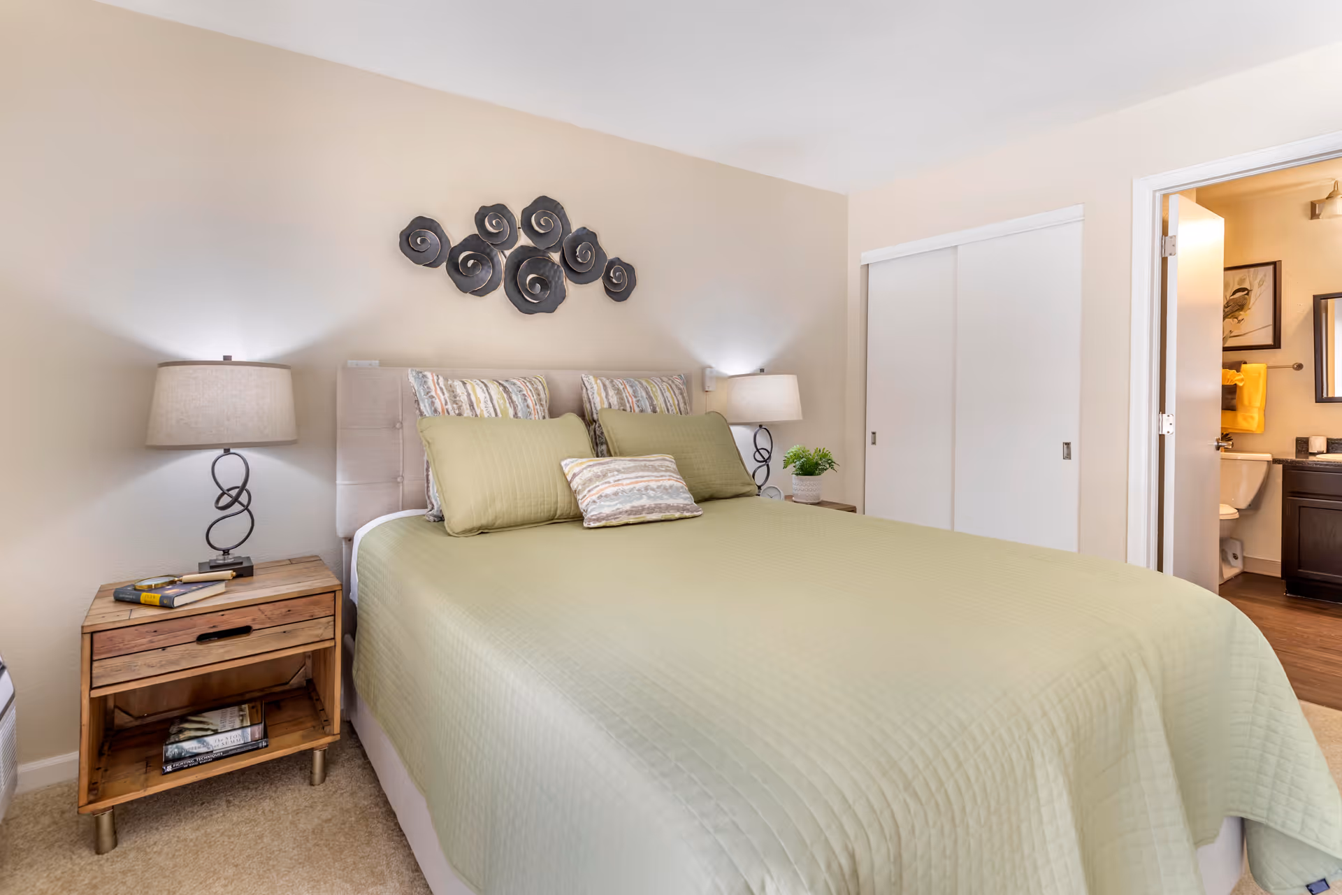 A bedroom with a neatly made bed covered in a light green quilt and matching pillows. There are two wooden nightstands on either side of the bed, each with a lamp. Above the bed is a decorative wall art piece with circular metal designs. To the right, there is a sliding closet door and an open door leading to a bathroom with visible sink, mirror, and towel.