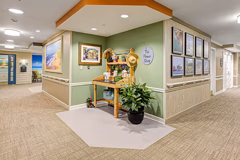 Interior hallway of a senior living facility with beige carpet and light green walls. A small wooden flower shop display table with gardening hats, pots, and plants is positioned in a corner under an orange ceiling accent. Framed pictures and artwork decorate the walls along the hallway.