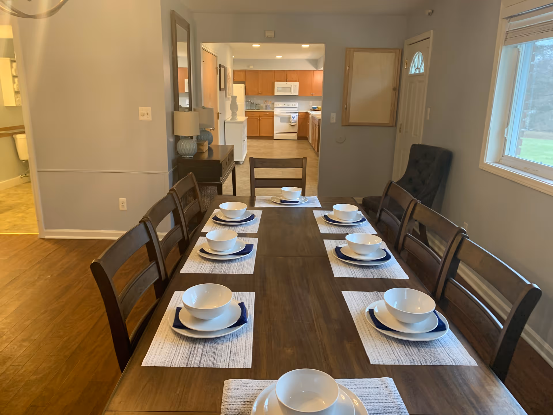 A dining room with a long wooden table set for six people with white bowls, plates, and blue napkins on white placemats. The room has wooden chairs around the table, a window on the right side, and a view into a kitchen with wooden cabinets and white appliances in the background.
