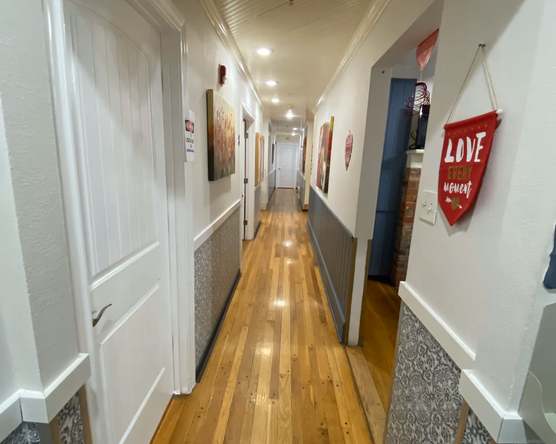 A long, narrow hallway with wooden floors and white walls, decorated with paintings and a red hanging sign that says 'LOVE EVERY moment'. Several white doors line the hallway, and recessed ceiling lights illuminate the space.