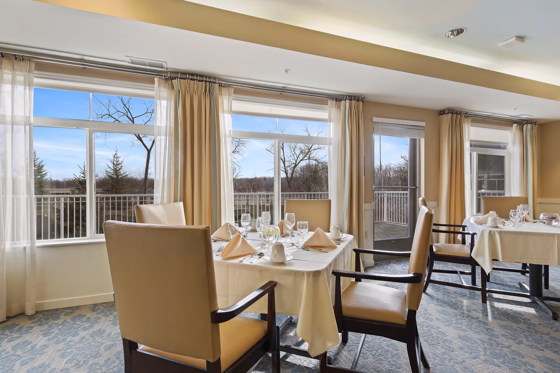 A bright dining room with large windows and glass doors letting in natural light. The room features tables set with white tablecloths, napkins, glassware, and silverware. Beige cushioned chairs surround the tables. Outside the windows, trees and a balcony railing are visible under a clear blue sky.