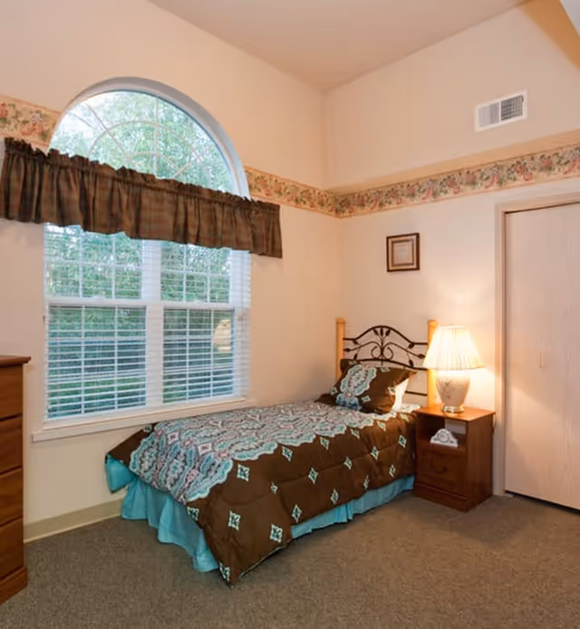 A cozy bedroom with a single bed covered in a brown and turquoise patterned comforter. The bed has a decorative metal headboard and is positioned next to a wooden nightstand with a lamp and a telephone. There is a large arched window with white blinds and a brown valance, allowing natural light to brighten the room. The walls are light-colored with a floral wallpaper border near the ceiling.