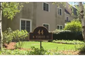 A black and gold sign in front of a beige building with multiple windows. The sign reads '44 W Webster St' and is surrounded by green grass, bushes, and trees.