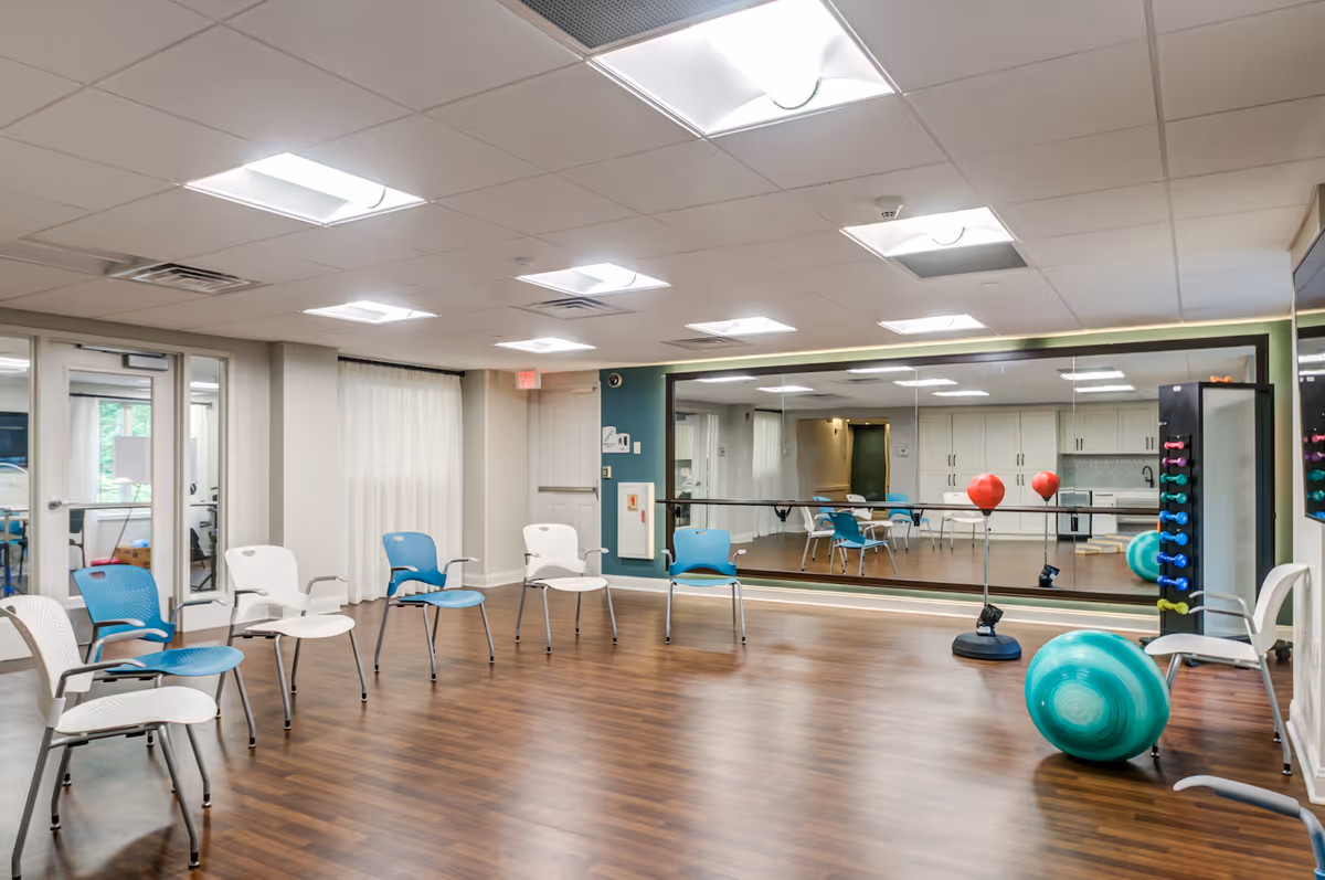 A spacious exercise room with wooden flooring, several white and blue chairs arranged in a semi-circle, a large wall mirror, exercise balls, and a rack of colorful dumbbells. The room has bright ceiling lights and large windows with sheer curtains.