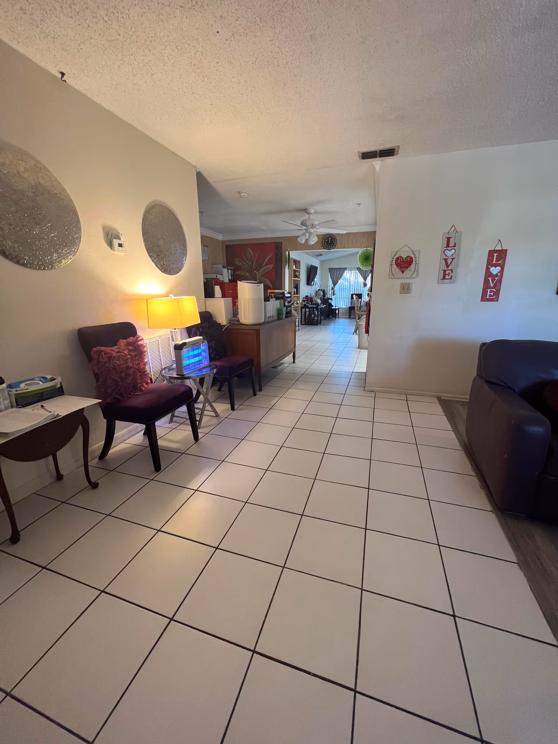 Interior view of a senior living facility hallway with white tiled floor, two purple chairs with red cushions, a small table with a lamp and a device emitting blue light. The hallway leads to a dining area with tables and chairs. The walls have decorative round mirrors and hanging signs with the word 'LOVE'.