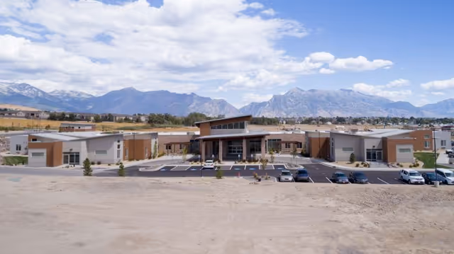 Wide exterior view of a modern senior living facility named Pointe Meadows with a large parking lot in front, surrounded by open land and mountains in the background under a partly cloudy sky.