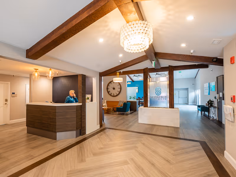 Reception area of Santa Cruz Post Acute facility with a receptionist sitting behind a modern wooden desk. The space features wooden ceiling beams, a large chandelier, a seating area with chairs and a clock on the wall, and a sign displaying the facility's name.