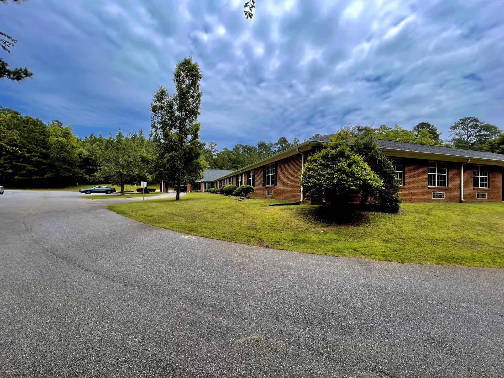 Low single-story brick memory care building with a lawn, trees and a parking lot under a cloudy sky.