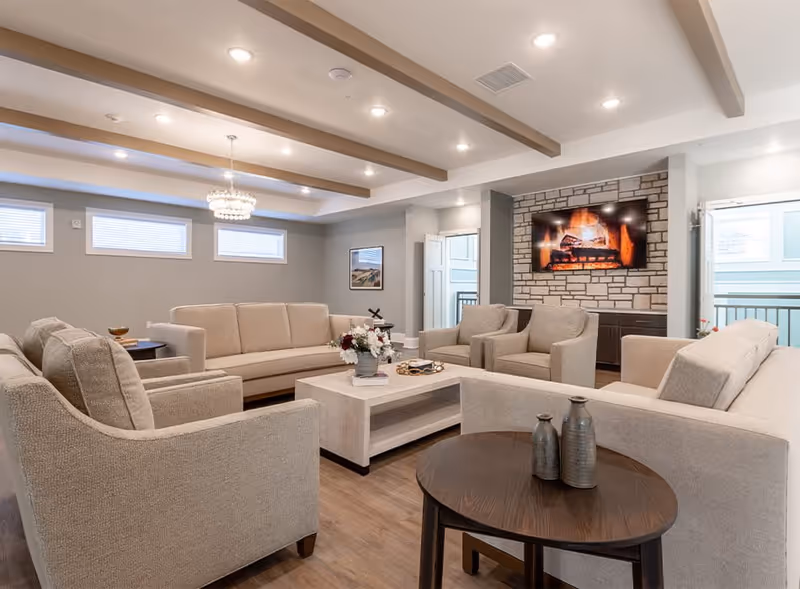 Spacious communal living room with beige sofas arranged around a central coffee table and a wall-mounted TV above a stone fireplace.