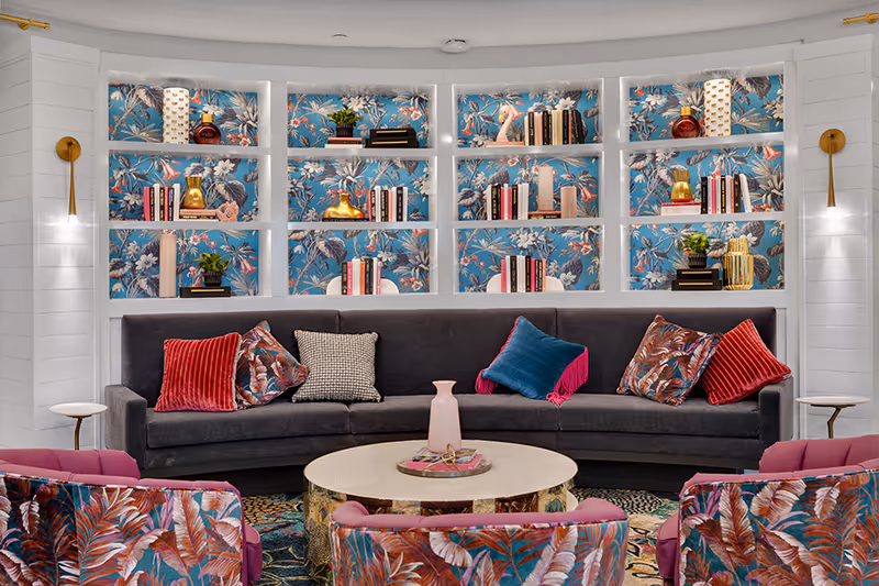 A stylish lounge with a dark gray sofa, colorful patterned pillows and chairs, a round coffee table, and built-in shelves backed with floral wallpaper and decorative items.
