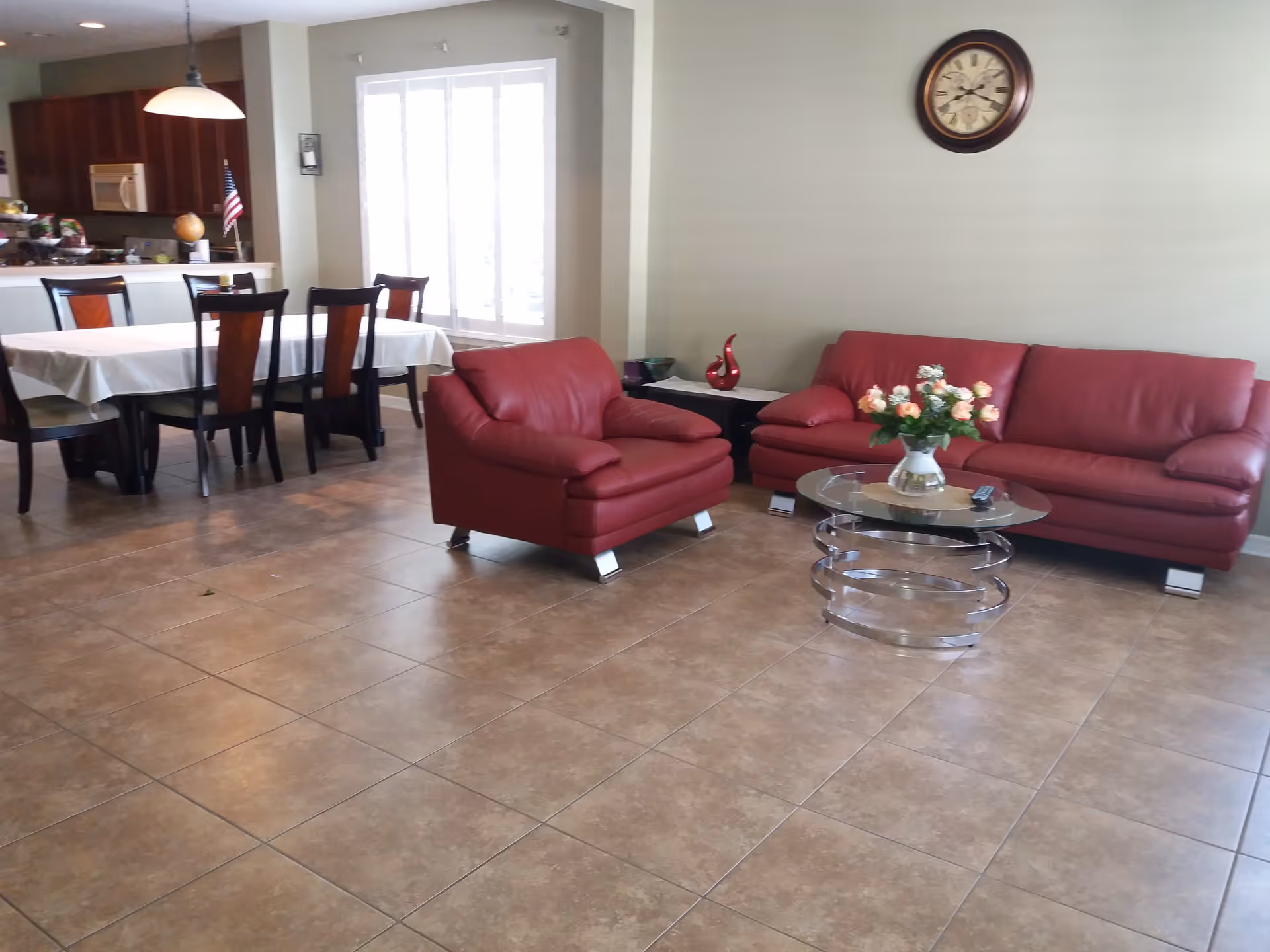 A spacious living area with a red leather sofa and matching armchair, a glass coffee table with a vase of flowers, a wall clock, and a dining area with a table covered with a white tablecloth and six chairs. The kitchen is visible in the background with wooden cabinets and a microwave.