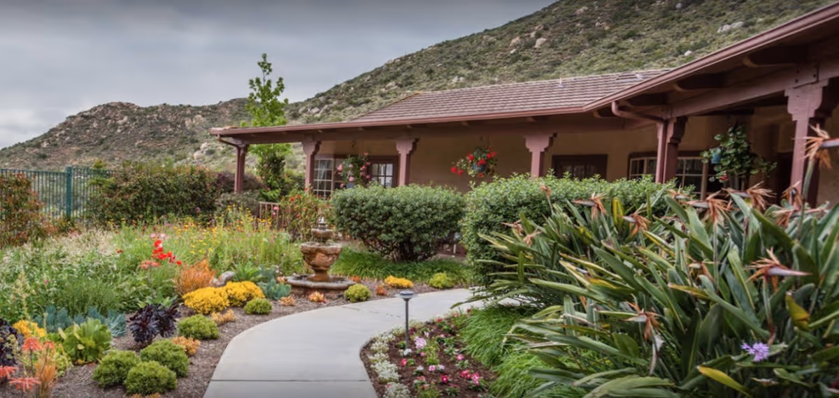 A garden pathway leading to a covered porch area of a building with hanging flower baskets and lush greenery, set against a backdrop of rocky hills under a cloudy sky.