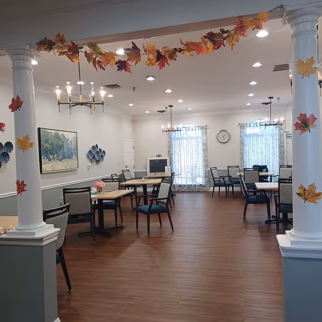 A spacious dining room decorated with autumn-themed garlands of colorful leaves hanging from the ceiling and wrapped around white columns. The room has wooden flooring, multiple tables with chairs arranged around them, and large windows with sheer curtains letting in natural light. The walls are adorned with a landscape painting and decorative plates, and a clock is mounted between the windows.