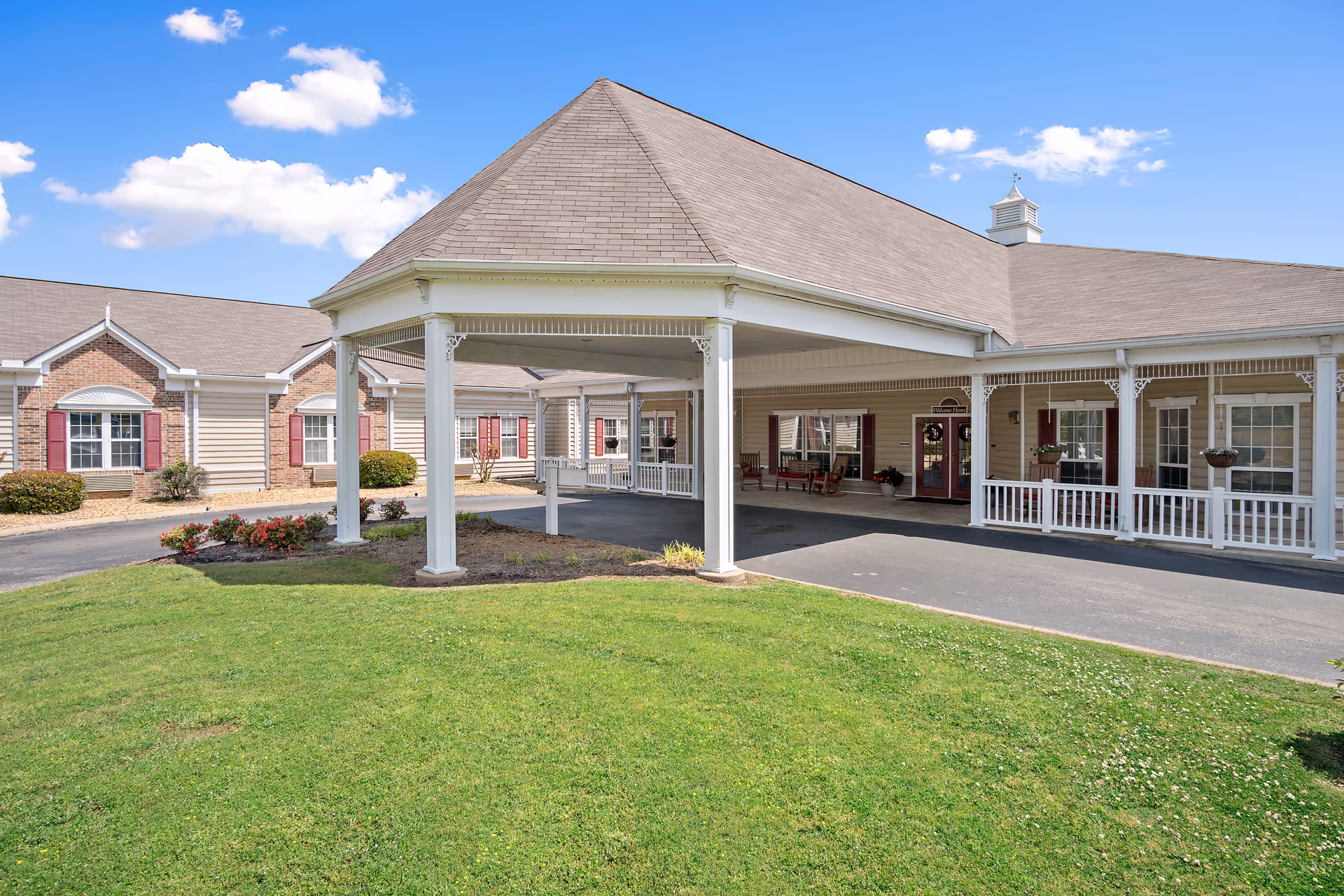 Exterior view of HarborChase of Jasper senior living facility showing a covered entrance with white pillars, a paved driveway, green lawn, and a building with beige siding, red shutters, and white trim under a blue sky with some clouds.