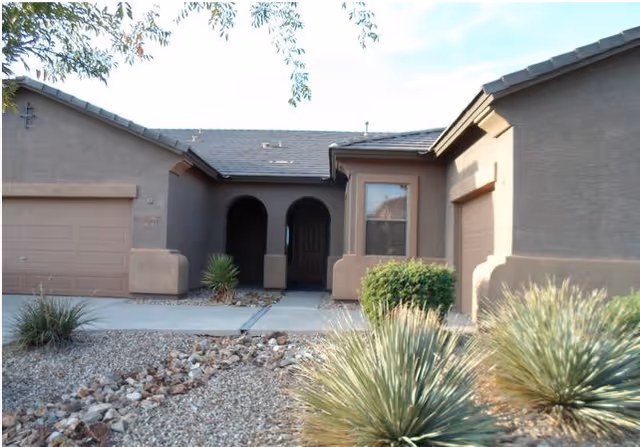 Front exterior view of a single-story house with a brown and beige color scheme, featuring two garage doors, a central entrance with an arched doorway, and desert landscaping with rocks and spiky plants.
