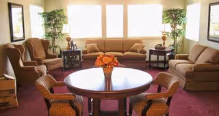 A cozy living room with a round wooden table and four chairs in the foreground, a beige sofa and two armchairs arranged around the room, two side tables with lamps, two tall potted plants near the windows, and framed artwork on the walls.