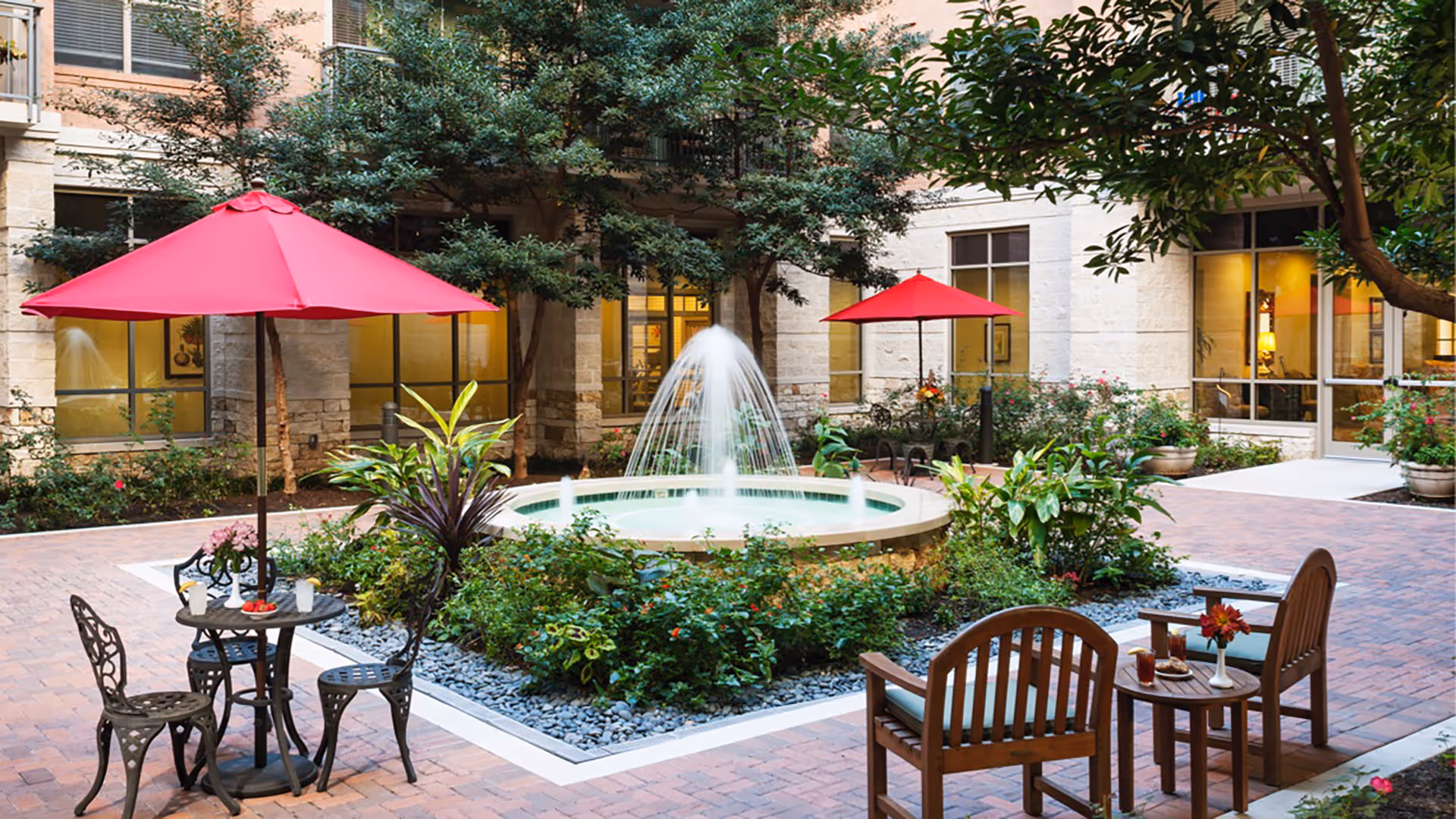 Outdoor courtyard area with a central water fountain surrounded by greenery and plants. There are two red umbrellas shading seating areas with metal and wooden chairs and small tables. The courtyard is paved with bricks and is adjacent to a building with large windows.