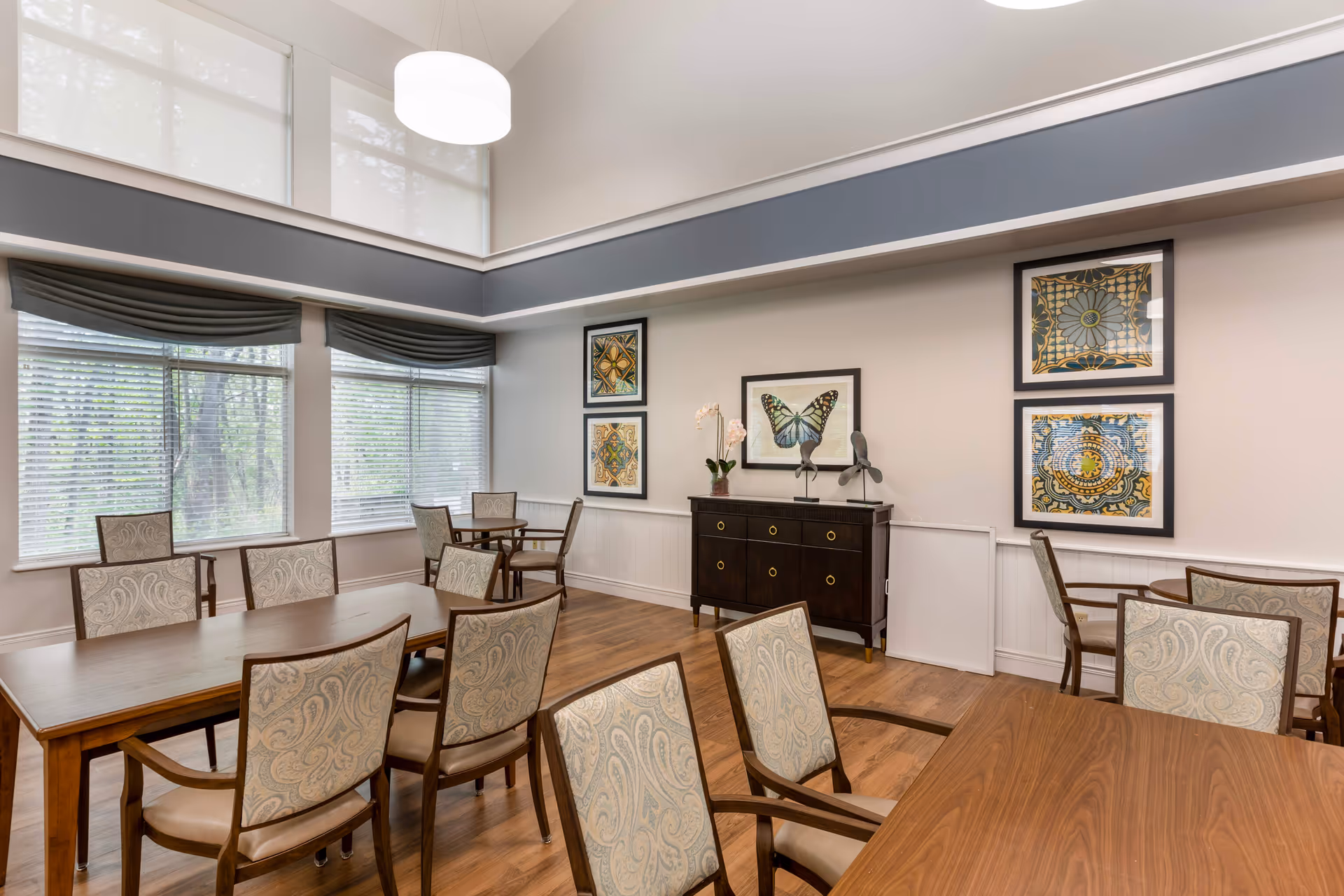 A bright dining room with multiple wooden tables and upholstered chairs arranged neatly. Large windows with blinds and valances allow natural light to fill the space. The walls are decorated with framed artwork including a butterfly and intricate patterns. A dark wooden sideboard with decorative items sits against one wall. The room has a high ceiling with a modern hanging light fixture.