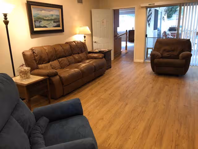A living room with wooden flooring featuring a brown leather sofa, a matching brown leather armchair, and a blue upholstered armchair. There are two side tables with lamps, a framed landscape painting on the wall, and a doorway leading to another room and a sliding glass door opening to an outdoor area.