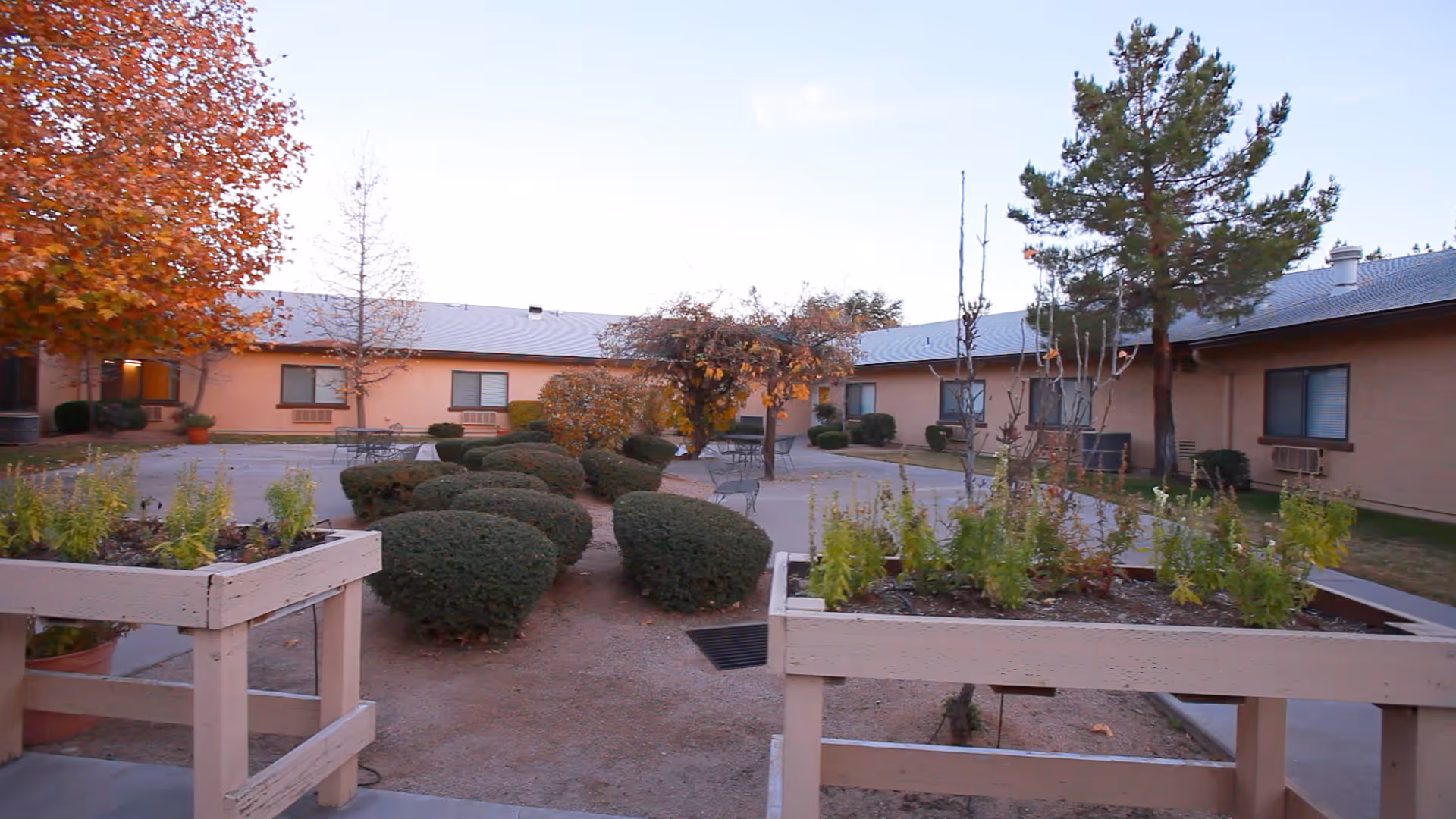Outdoor courtyard area at Payson Care Center with trimmed bushes, small trees, raised garden beds, and patio tables and chairs surrounded by a single-story building under a clear sky.