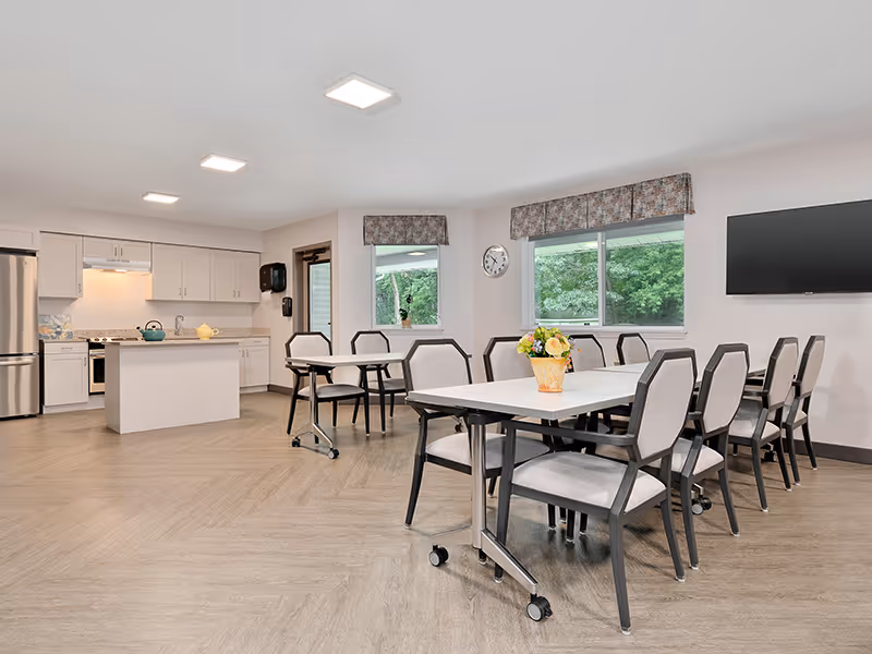 A bright and clean dining area in a senior living facility featuring two tables with cushioned chairs on wheels. A vase with flowers is placed on the table in the foreground. The room has large windows with patterned valances, a wall clock, a flat-screen TV mounted on the wall, and a kitchen area with white cabinets, a refrigerator, stove, and an island in the background.