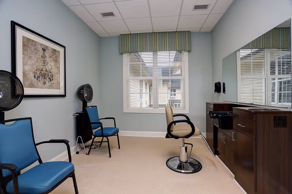A small salon room with a beige salon chair in front of a large mirror and dark wooden cabinetry. Two blue waiting chairs are positioned against the left wall, which also features a framed artwork. A window with white blinds and a green patterned valance lets in natural light.