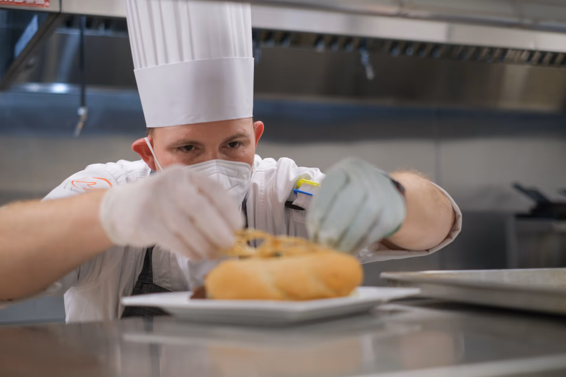 A chef wearing a white chef hat, white gloves, and a face mask is carefully placing garnish on a plated sandwich in a professional kitchen.