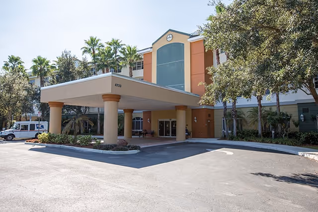 Entrance of a multi-story senior living facility with a covered porte-cochère, columns, landscaping, and a driveway.