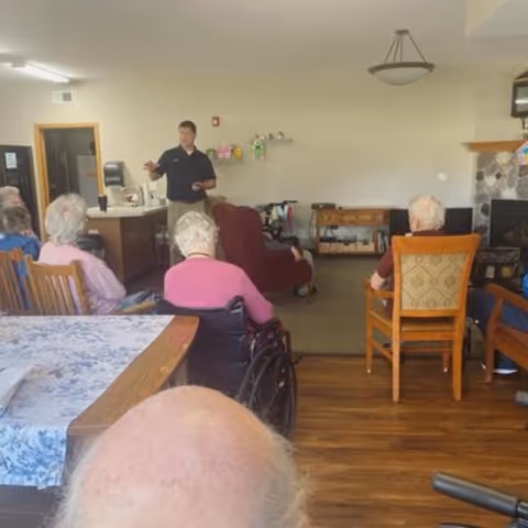 Several elderly residents seated in chairs and wheelchairs facing a standing staff member who is speaking in a communal living room of an assisted-living facility.