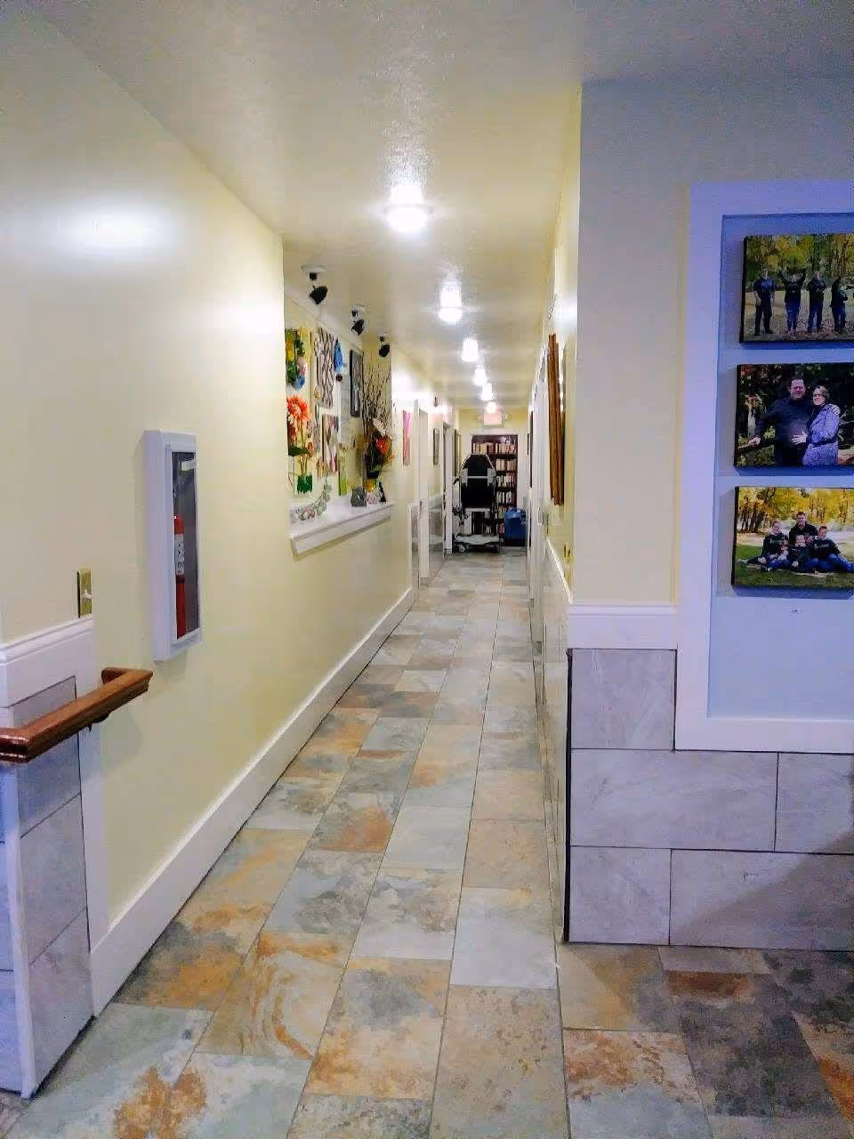 A long, well-lit hallway with tiled flooring and cream-colored walls. The left wall features a fire extinguisher cabinet and a display of colorful artwork. The right side has framed family photos mounted on the wall. At the end of the hallway, there is a bookshelf and a chair.