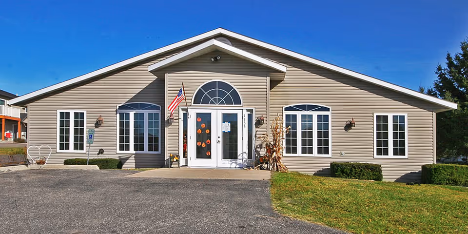 Front exterior of a single-story beige senior living building with a central glass entrance, American flag, and paved driveway.