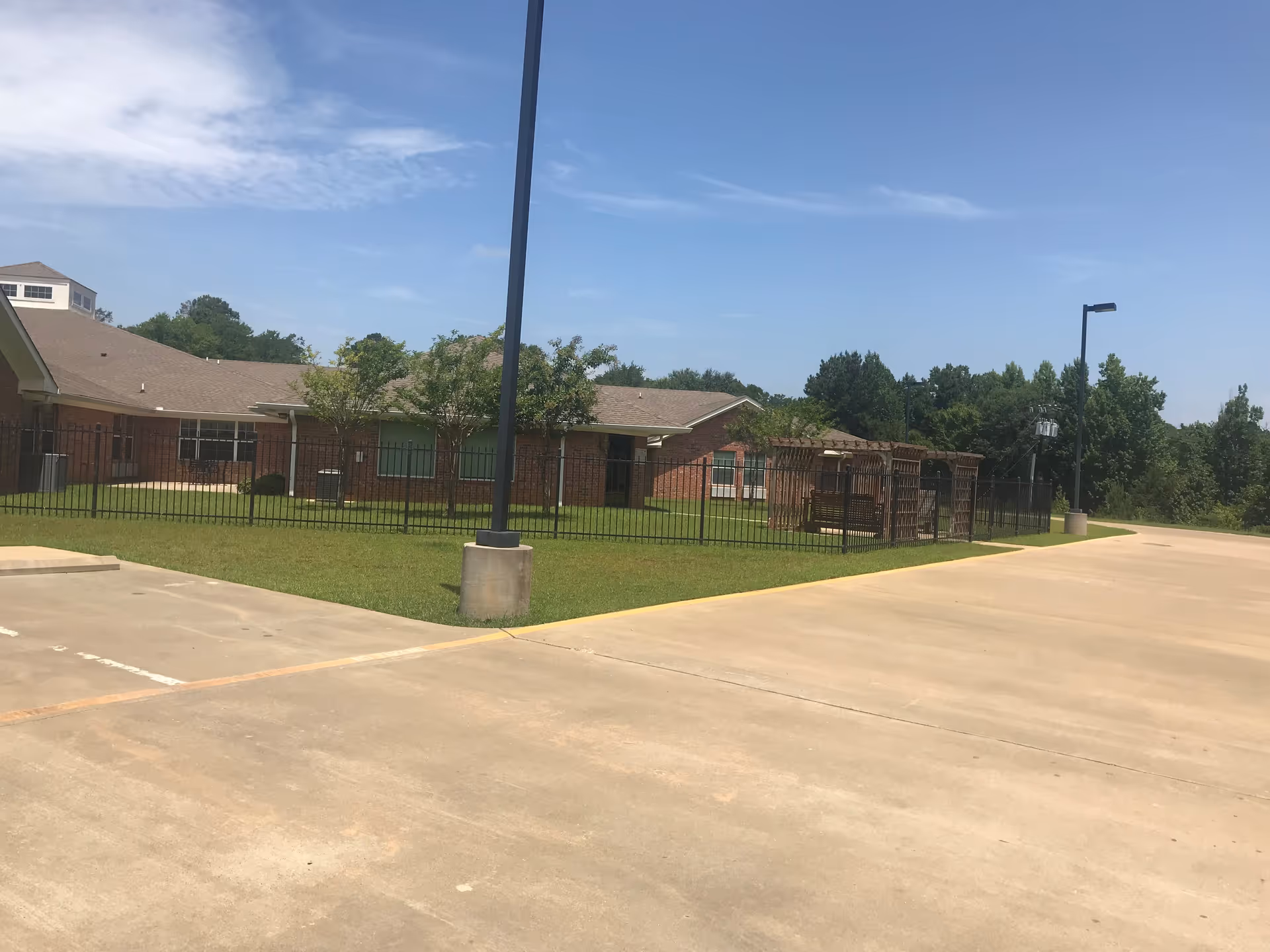 Outdoor view of The Arbors Healthcare and Rehabilitation Center showing a brick building with a fenced grassy area, trees, and a paved parking lot under a partly cloudy sky.