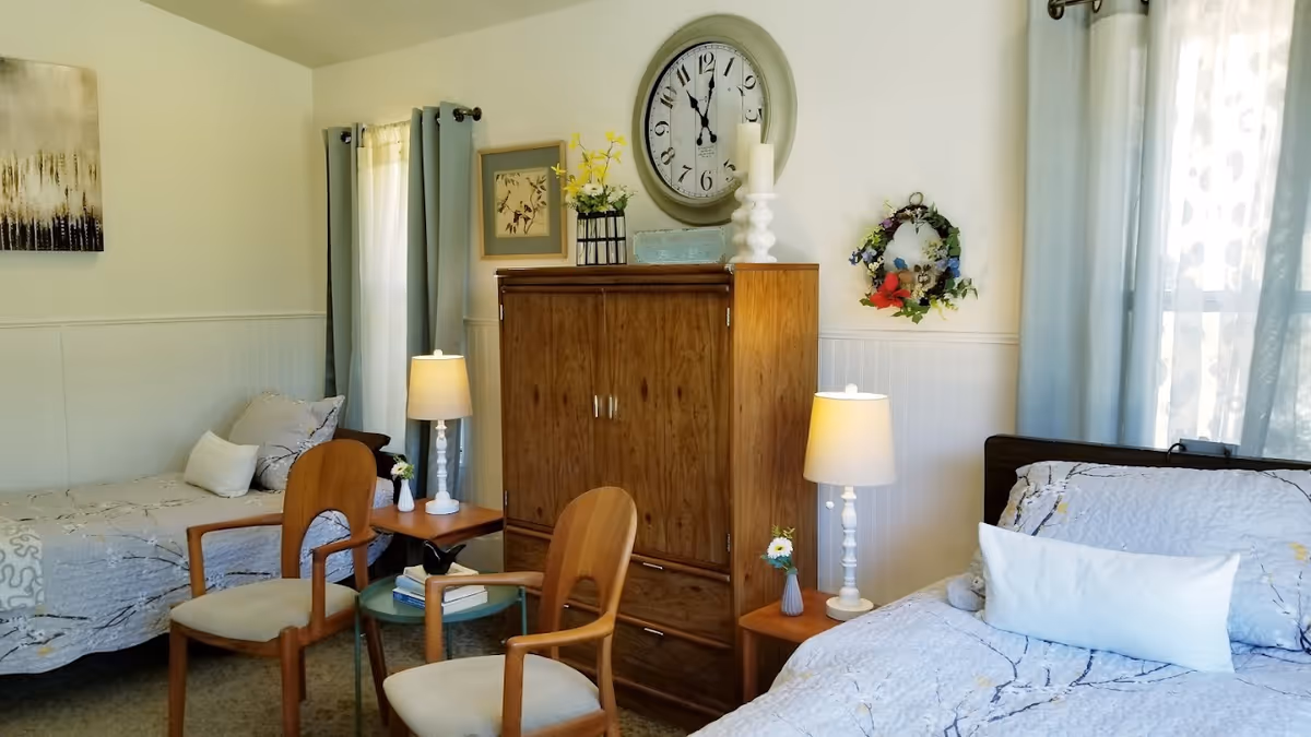 A cozy eldercare bedroom with two beds, each with light-colored bedding and pillows. Between the beds is a wooden cabinet with a large round clock above it, flanked by two table lamps on small wooden side tables. Two wooden chairs with cushions are positioned around a small glass-top table with books. The room has light-colored walls, curtains on the windows, and decorative wall art including a floral wreath.