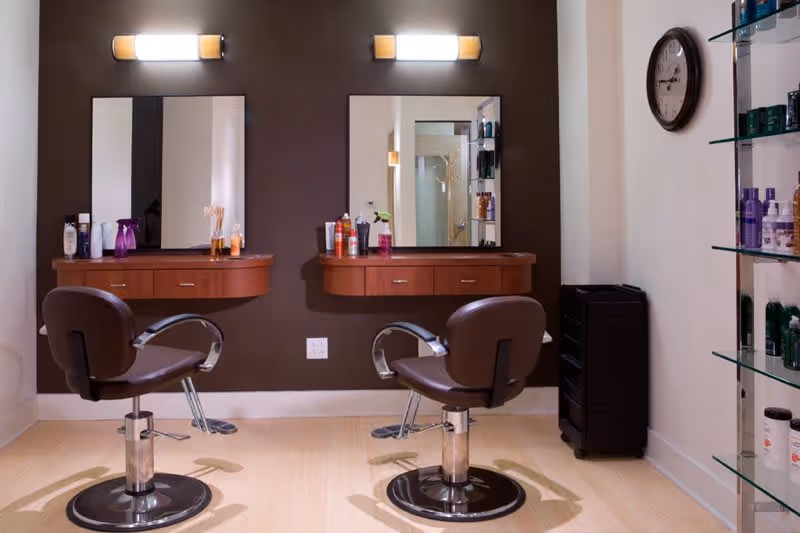 Interior view of a salon area with two brown styling chairs in front of two mirrors mounted on a dark brown wall. Each mirror has a wooden shelf below it holding various hair care products. To the right, there is a glass shelving unit with more hair products and a wall clock above it.
