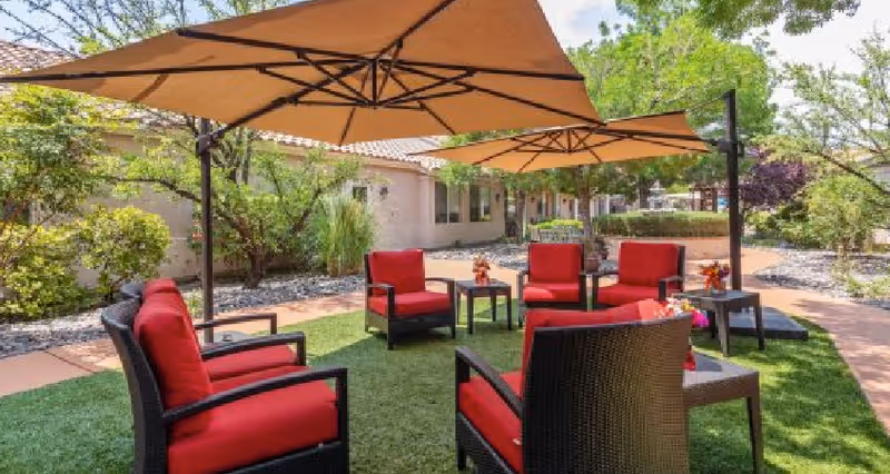 Outdoor seating area with six black wicker chairs featuring red cushions arranged in a circle on green artificial grass. Two large beige patio umbrellas provide shade. The area is surrounded by trees, shrubs, and a building with windows in the background.