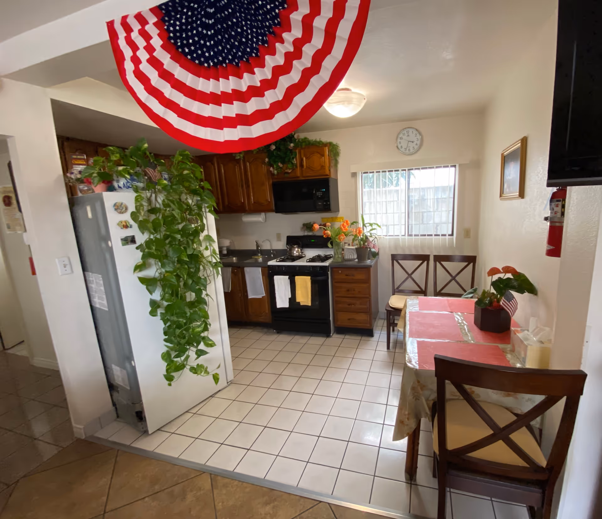 A small kitchen and dining area with white tiled floor and wooden cabinets. There is a white refrigerator with a large green plant hanging on its side. A black stove and microwave are against the far wall under a window with vertical blinds. A round clock is mounted above the window. A dining table with a floral tablecloth and red placemats is set with three wooden chairs. An American flag-themed decoration hangs from the ceiling.