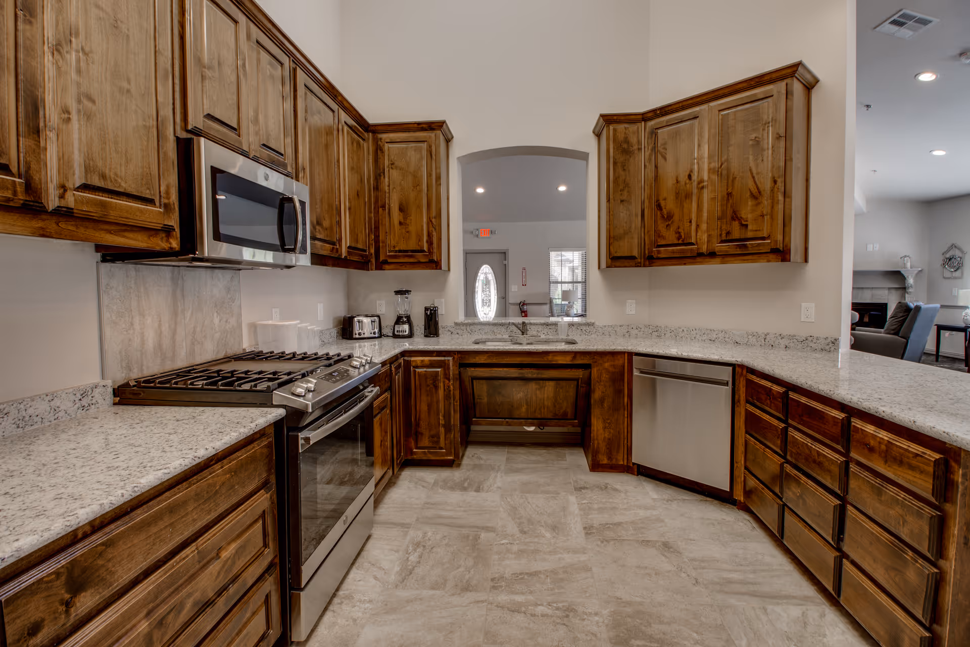 A modern kitchen with wooden cabinets, granite countertops, a stainless steel gas stove with oven, microwave, dishwasher, and small appliances like a toaster and blender. The kitchen has a tiled floor and an open archway leading to a living area with a fireplace and seating.