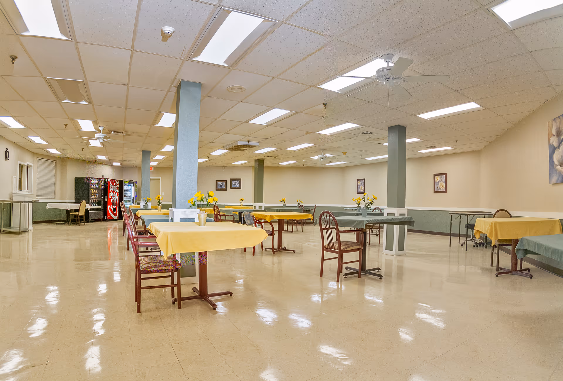 A spacious dining room with several tables covered in yellow and green tablecloths, each decorated with small flower arrangements. There are wooden chairs around the tables, vending machines in the background, and fluorescent ceiling lights illuminating the room.
