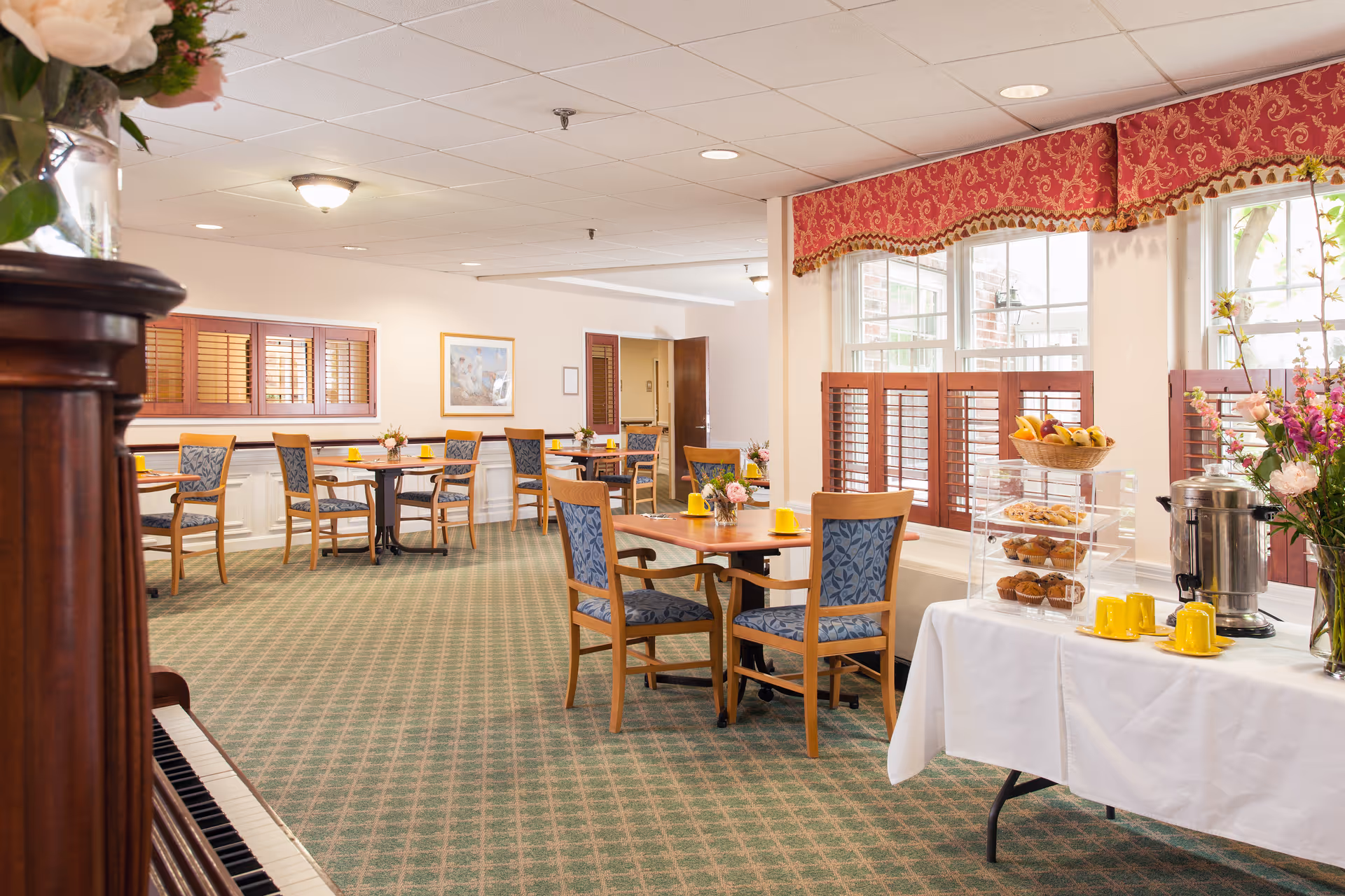 A bright dining room in a senior living facility with several wooden tables and chairs arranged neatly. Each table has a small flower arrangement and yellow cups. There is a table on the right side with a white tablecloth holding a coffee urn, yellow cups, and a tiered tray with muffins and fruit. Large windows with red patterned valances allow natural light to fill the room.
