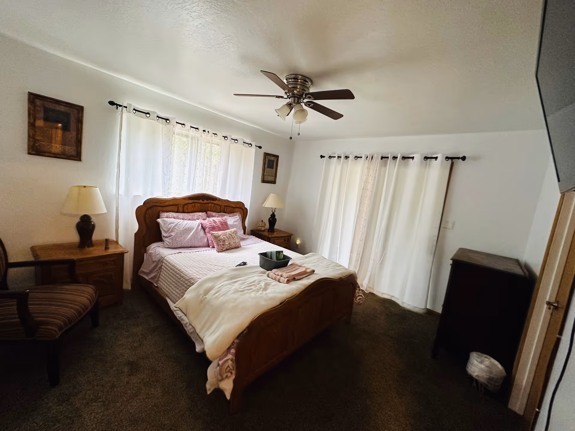 Sunlit bedroom with a wooden bed, bedside tables and lamps, ceiling fan, and white curtains.